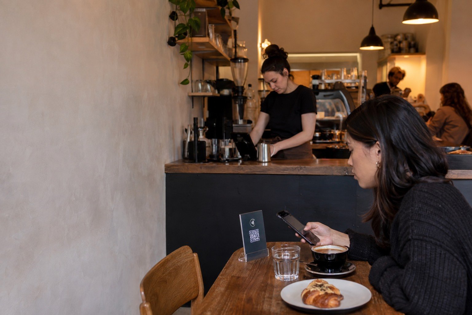 A café counter with a card reader, and a guest at a nearby table ordering by tapping the display