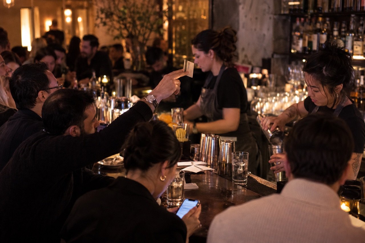 Guests three deep at a busy bar, one raising a hand to get the bartender's attention