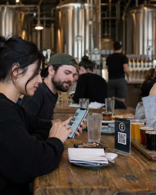 A guest orders from her phone at a long table in a craft brewery taproom